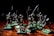 A group of miniature skeleton figures is positioned on a wooden surface. These figures are intricately detailed, each holding various types of weapons such as swords and spears. They stand on green square bases, giving them a battle-ready appearance. The lighting creates dramatic shadows, emphasizing the eerie and menacing nature of the scene.