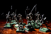A group of miniature skeleton figures is positioned on a wooden surface. These figures are intricately detailed, each holding various types of weapons such as swords and spears. They stand on green square bases, giving them a battle-ready appearance. The lighting creates dramatic shadows, emphasizing the eerie and menacing nature of the scene.