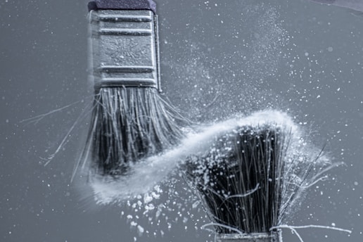 Two paintbrushes in motion, capturing the dynamic energy of white powder or dust dispersed between them against a grey background.