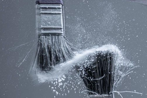 Two paintbrushes in motion, capturing the dynamic energy of white powder or dust dispersed between them against a grey background.