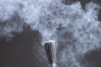 A close-up of a brush dispersing fine white powder into the air, creating a cloud of particles that swirl and float against a dark background. The texture and movement of the powder are captured with intricate detail.