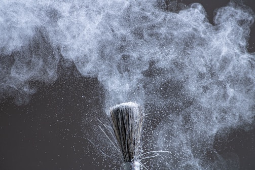 A close-up of a brush dispersing fine white powder into the air, creating a cloud of particles that swirl and float against a dark background. The texture and movement of the powder are captured with intricate detail.