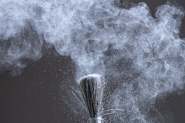 A close-up of a brush dispersing fine white powder into the air, creating a cloud of particles that swirl and float against a dark background. The texture and movement of the powder are captured with intricate detail.