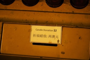 A close-up view of a donation box with a sign in both English and Chinese that reads 'Candle Donation $2'. The sign is placed on a mustard-yellow colored surface.