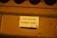 A close-up view of a donation box with a sign in both English and Chinese that reads 'Candle Donation $2'. The sign is placed on a mustard-yellow colored surface.