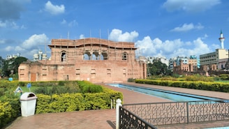 A historical brick structure under renovation with scaffolding stands prominently. Surrounding the building is a well-manicured garden with neatly trimmed bushes and a pathway lined with hedges. A long, narrow water feature runs parallel to the path. In the background, modern buildings and a tower with a pointed top are visible under a partly cloudy blue sky.