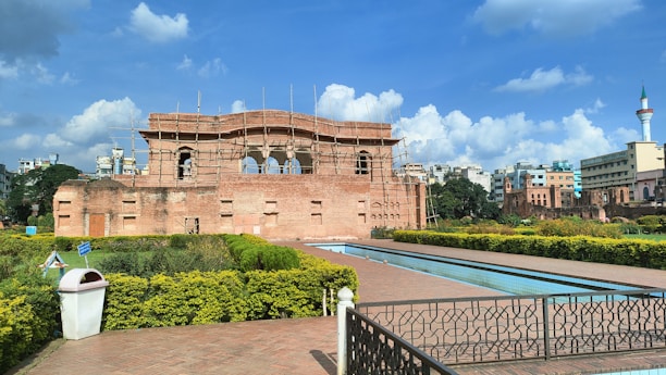 A historical brick structure under renovation with scaffolding stands prominently. Surrounding the building is a well-manicured garden with neatly trimmed bushes and a pathway lined with hedges. A long, narrow water feature runs parallel to the path. In the background, modern buildings and a tower with a pointed top are visible under a partly cloudy blue sky.