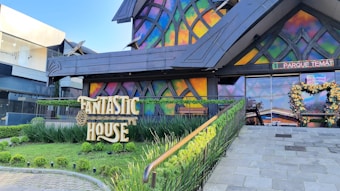 A colorful building facade with a stained-glass appearance, displaying the words 'Fantastic House' prominently. Neatly trimmed greenery frames the entrance, and a floral heart-shaped decoration is positioned near the doorway.