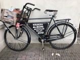 A vintage black bicycle is parked against a wall, featuring a wooden crate attached to the front. The crate contains metal containers and is secured with a red cloth. The bicycle has a large metal sign on the frame with text related to a store or service. The pavement consists of small, rectangular bricks.