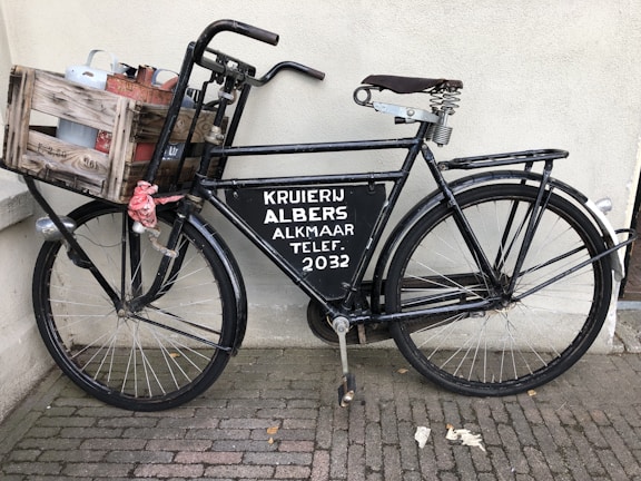 A vintage black bicycle is parked against a wall, featuring a wooden crate attached to the front. The crate contains metal containers and is secured with a red cloth. The bicycle has a large metal sign on the frame with text related to a store or service. The pavement consists of small, rectangular bricks.