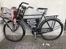 A vintage black bicycle is parked against a wall, featuring a wooden crate attached to the front. The crate contains metal containers and is secured with a red cloth. The bicycle has a large metal sign on the frame with text related to a store or service. The pavement consists of small, rectangular bricks.