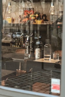 A cozy café setting with wooden furniture, glassware, and bottles visible through a window. Hanging light bulbs provide soft lighting. The background includes a shelf stocked with jars and fresh fruits.