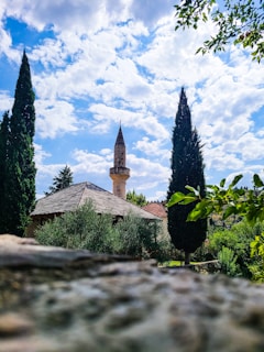 A picturesque scene depicting a mosque with a prominent minaret surrounded by lush greenery and tall cypress trees. The sky is partly cloudy, allowing patches of blue to peek through, enhancing the serene atmosphere. The foreground is slightly blurred, offering a sense of depth to the image.