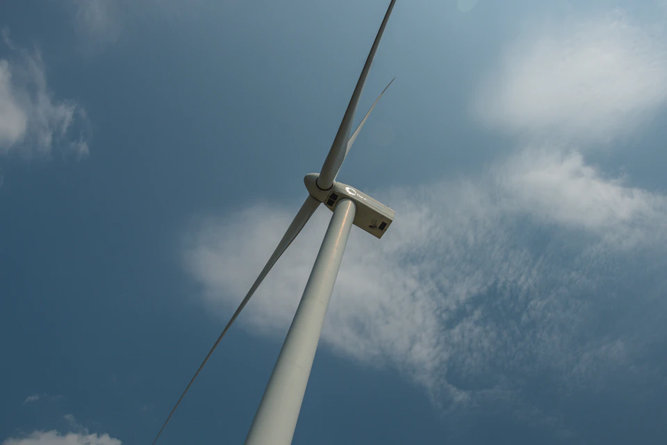 The image shows a large white wind turbine with four blades, standing tall against a clear blue sky dotted with fluffy white clouds. The perspective is from below, looking up at the towering structure and emphasizing its height and grandeur.