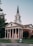 A red-brick church with white columns and a tall steeple topped with a cross stands prominently. The building features classic architectural design elements such as a triangular pediment and a symmetrical facade. The entrance includes three sets of red double doors. Surrounding the church are well-maintained lawns and a few trees. A sign on the lawn indicates an orientation event nearby.