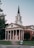A red-brick church with white columns and a tall steeple topped with a cross stands prominently. The building features classic architectural design elements such as a triangular pediment and a symmetrical facade. The entrance includes three sets of red double doors. Surrounding the church are well-maintained lawns and a few trees. A sign on the lawn indicates an orientation event nearby.