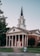 A red-brick church with white columns and a tall steeple topped with a cross stands prominently. The building features classic architectural design elements such as a triangular pediment and a symmetrical facade. The entrance includes three sets of red double doors. Surrounding the church are well-maintained lawns and a few trees. A sign on the lawn indicates an orientation event nearby.