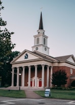 A red-brick church with white columns and a tall steeple topped with a cross stands prominently. The building features classic architectural design elements such as a triangular pediment and a symmetrical facade. The entrance includes three sets of red double doors. Surrounding the church are well-maintained lawns and a few trees. A sign on the lawn indicates an orientation event nearby.