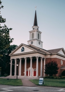 A red-brick church with white columns and a tall steeple topped with a cross stands prominently. The building features classic architectural design elements such as a triangular pediment and a symmetrical facade. The entrance includes three sets of red double doors. Surrounding the church are well-maintained lawns and a few trees. A sign on the lawn indicates an orientation event nearby.