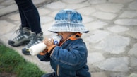 A child wearing a denim jacket and a matching bucket hat is drinking from a bottle. The scene takes place outdoors on a stone path, with grass visible in the bottom left. An adult's legs, wearing dark pants and hiking boots, are partially visible.