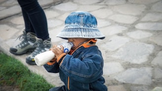 A child wearing a denim jacket and a matching bucket hat is drinking from a bottle. The scene takes place outdoors on a stone path, with grass visible in the bottom left. An adult's legs, wearing dark pants and hiking boots, are partially visible.