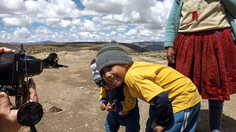 Two children are playfully posing next to an adult, who is partially visible. One child is smiling while wearing a gray beanie and yellow shirt. A camera is held by a photographer on the left capturing the moment, and a black dog is lying on the ground in the background. The setting is an open, rocky landscape under a partly cloudy sky.