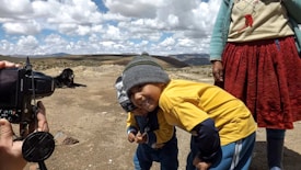 Two children are playfully posing next to an adult, who is partially visible. One child is smiling while wearing a gray beanie and yellow shirt. A camera is held by a photographer on the left capturing the moment, and a black dog is lying on the ground in the background. The setting is an open, rocky landscape under a partly cloudy sky.