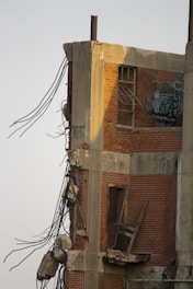 A partially demolished brick building with exposed concrete beams and steel rebar jutting out. The structure's interior is visible, including broken windows and graffiti on the remaining wall. Debris is scattered around the damaged section.