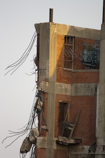 A partially demolished brick building with exposed concrete beams and steel rebar jutting out. The structure's interior is visible, including broken windows and graffiti on the remaining wall. Debris is scattered around the damaged section.