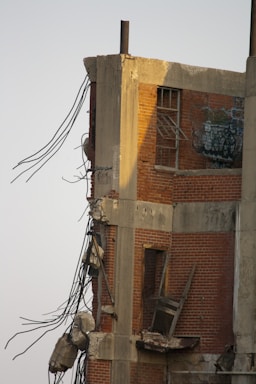 A partially demolished brick building with exposed concrete beams and steel rebar jutting out. The structure's interior is visible, including broken windows and graffiti on the remaining wall. Debris is scattered around the damaged section.