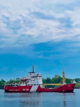A large red and white Canadian Coast Guard ship is stationed on calm waters. The sky above is mostly cloudy with patches of blue peeking through. In the background, there's a stretch of green foliage and trees, possibly indicating a shoreline or island.