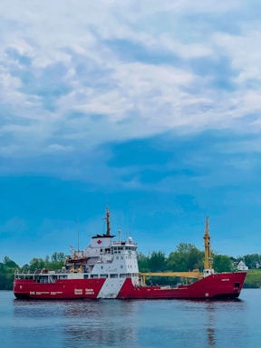 A large red and white Canadian Coast Guard ship is stationed on calm waters. The sky above is mostly cloudy with patches of blue peeking through. In the background, there's a stretch of green foliage and trees, possibly indicating a shoreline or island.