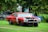 A red vintage car with visible rust and missing front parts is parked on a grassy lawn. The car appears to be an old model undergoing restoration or used for scrap. In the background, there are trees and another vehicle partially visible.