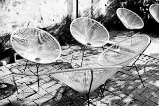 A group of modern, circular chairs with woven seats is arranged on a stone-paved outdoor patio. The chairs have thin, black metal legs and the seats are constructed from numerous cords creating a radial pattern. There is a rough concrete wall in the background, and some shadow patterns are cast on the ground from the sunlight.