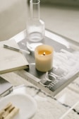 A minimalist coffee table setup featuring a lit candle, a glass carafe, and a couple of books, one of which has the title 'Dior Couture'. The arrangement includes a pen and some wooden incense sticks on a small tray, all on a clear glass surface.