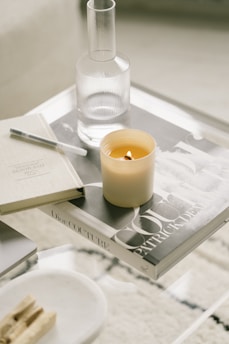 A minimalist coffee table setup featuring a lit candle, a glass carafe, and a couple of books, one of which has the title 'Dior Couture'. The arrangement includes a pen and some wooden incense sticks on a small tray, all on a clear glass surface.