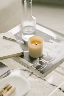 A minimalist coffee table setup featuring a lit candle, a glass carafe, and a couple of books, one of which has the title 'Dior Couture'. The arrangement includes a pen and some wooden incense sticks on a small tray, all on a clear glass surface.