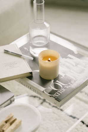 A minimalist coffee table setup featuring a lit candle, a glass carafe, and a couple of books, one of which has the title 'Dior Couture'. The arrangement includes a pen and some wooden incense sticks on a small tray, all on a clear glass surface.
