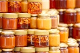 A collection of glass jars filled with various shades of honey are neatly arranged in stacks. Some jars have golden lids and contain suspended nuts within the amber liquid. The assortment displays a range of golden hues, from light to dark amber.