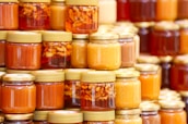 A collection of glass jars filled with various shades of honey are neatly arranged in stacks. Some jars have golden lids and contain suspended nuts within the amber liquid. The assortment displays a range of golden hues, from light to dark amber.