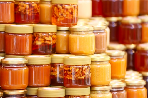 A collection of glass jars filled with various shades of honey are neatly arranged in stacks. Some jars have golden lids and contain suspended nuts within the amber liquid. The assortment displays a range of golden hues, from light to dark amber.