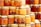 A collection of glass jars filled with various shades of honey are neatly arranged in stacks. Some jars have golden lids and contain suspended nuts within the amber liquid. The assortment displays a range of golden hues, from light to dark amber.