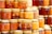 A collection of glass jars filled with various shades of honey are neatly arranged in stacks. Some jars have golden lids and contain suspended nuts within the amber liquid. The assortment displays a range of golden hues, from light to dark amber.