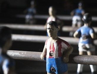 Several foosball table figures stand in focus, with their red and blue striped shirts, positioned on metal rods, casting shadows. The lighting creates a dramatic contrast, highlighting the figures against a dark background.