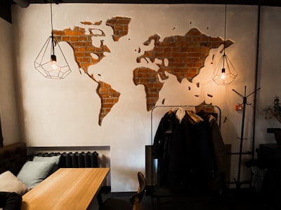 A cozy indoor space with a wooden table, cushions on a bench, and a world map mural made of exposed bricks on a white wall. Two modern pendant lights with geometric wire frames hang from the ceiling. A coat rack with multiple jackets is visible next to the table.