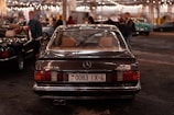 A classic Mercedes-Benz 280 SE with AMG branding is parked indoors, captured from the rear view. The license plate is visible, surrounded by a dimly lit environment filled with other people and vintage cars. Overhead string lights add a warm ambiance to the scene.