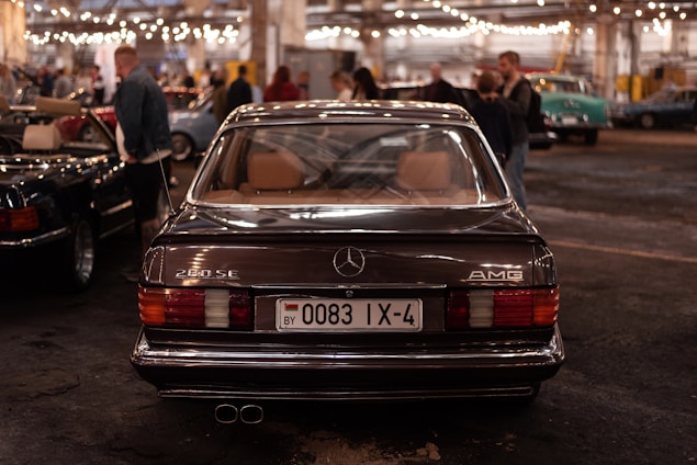 A classic Mercedes-Benz 280 SE with AMG branding is parked indoors, captured from the rear view. The license plate is visible, surrounded by a dimly lit environment filled with other people and vintage cars. Overhead string lights add a warm ambiance to the scene.