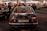 A classic Mercedes-Benz 280 SE with AMG branding is parked indoors, captured from the rear view. The license plate is visible, surrounded by a dimly lit environment filled with other people and vintage cars. Overhead string lights add a warm ambiance to the scene.