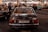 A classic Mercedes-Benz 280 SE with AMG branding is parked indoors, captured from the rear view. The license plate is visible, surrounded by a dimly lit environment filled with other people and vintage cars. Overhead string lights add a warm ambiance to the scene.
