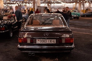 A classic Mercedes-Benz 280 SE with AMG branding is parked indoors, captured from the rear view. The license plate is visible, surrounded by a dimly lit environment filled with other people and vintage cars. Overhead string lights add a warm ambiance to the scene.